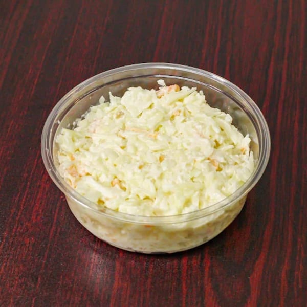 A clear plastic container filled with creamy coleslaw, featuring shredded cabbage and carrots, on a dark wooden surface.