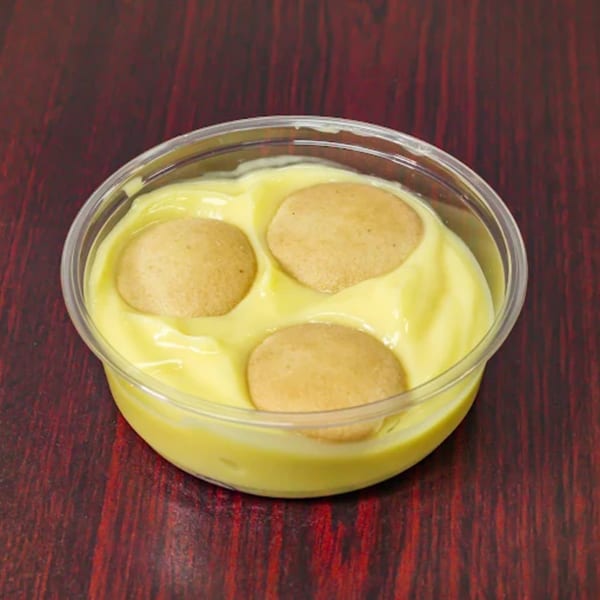 Banana pudding with three vanilla wafers in a clear plastic cup on a dark red table.