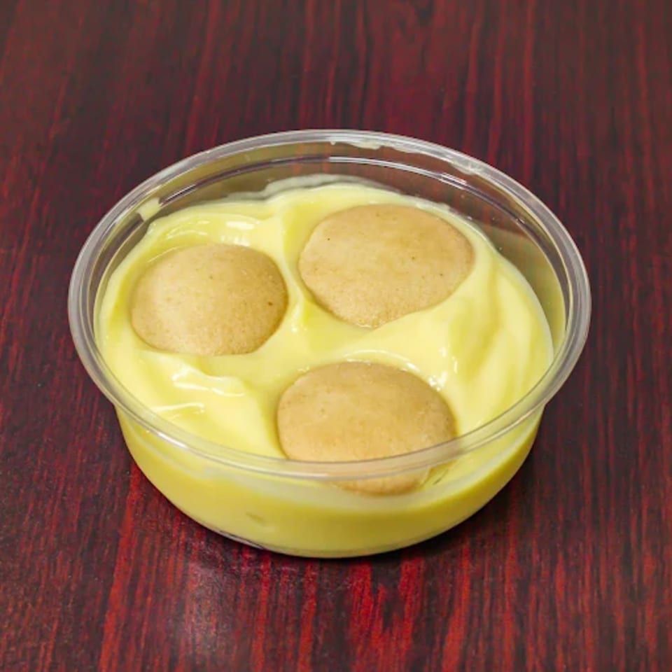 Banana pudding with three vanilla wafers in a clear plastic cup on a dark red table.