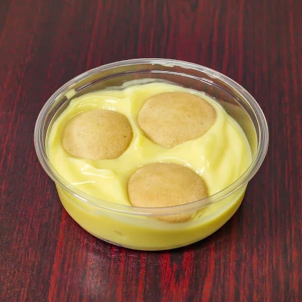 A clear plastic cup of banana pudding with three vanilla wafers on a dark red wooden surface.