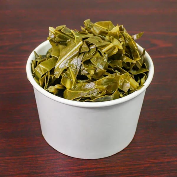 A white paper cup filled with cooked collard greens sits on a dark wooden surface.