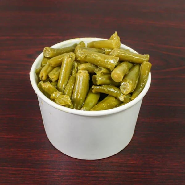 A white paper cup filled with cooked green beans, sitting on a dark wooden surface.