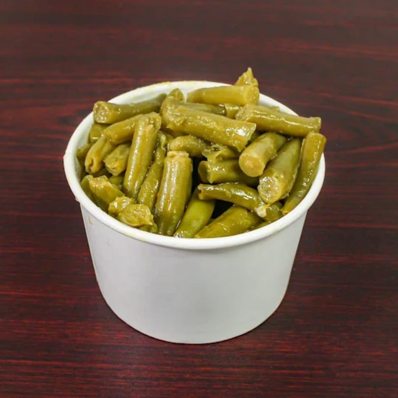 A white paper cup filled with cooked green beans, sitting on a dark wooden surface.